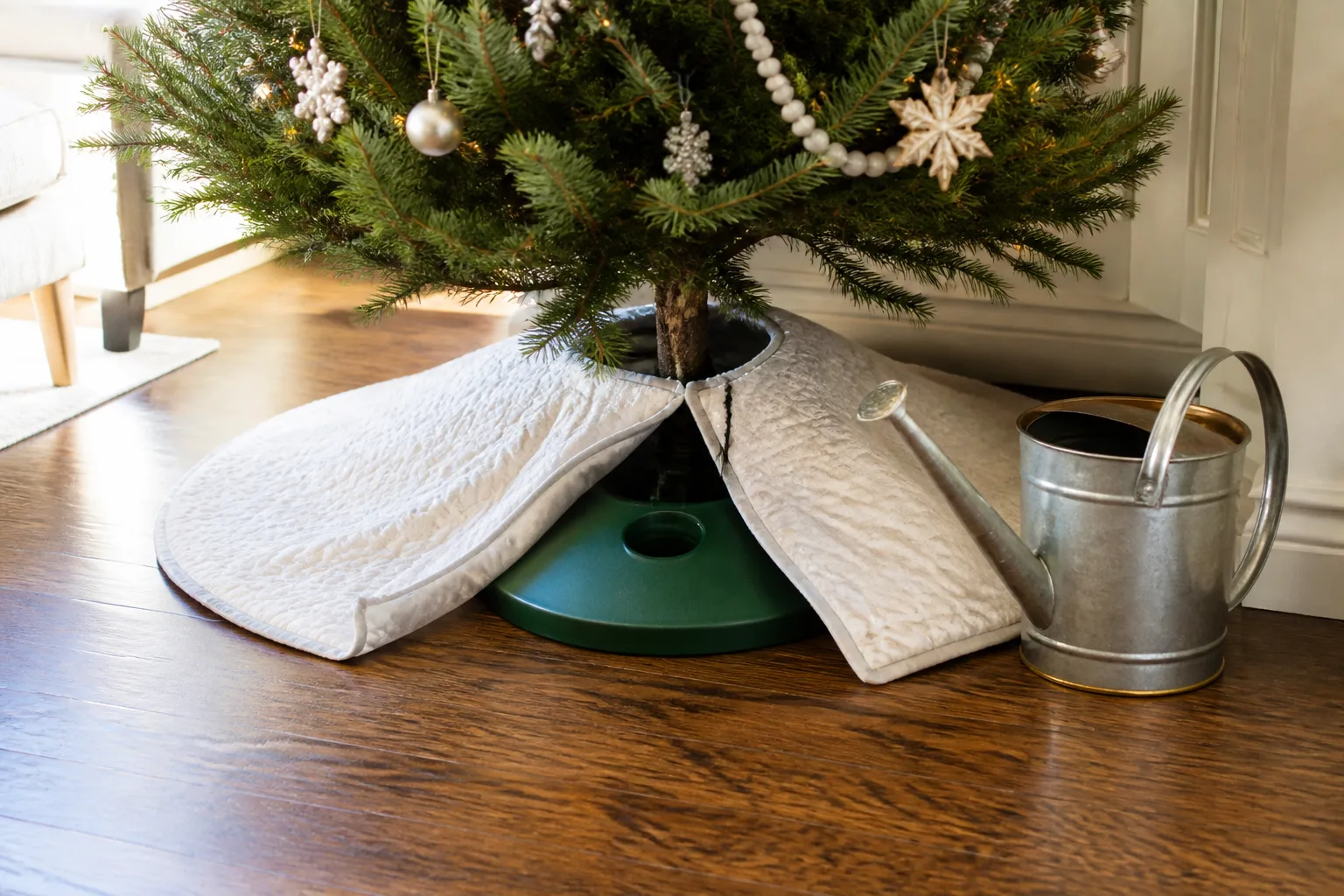 Christmas tree skirt split open toward a water-holding stand with a long-spout watering can nearby