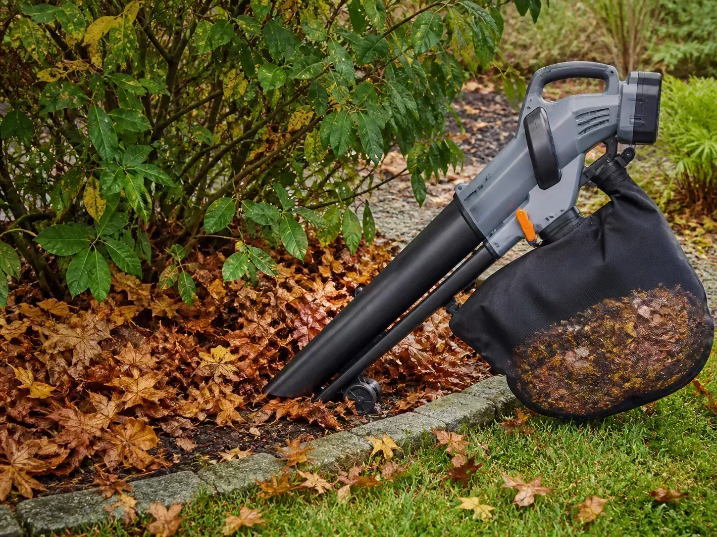 A generic handheld cordless leaf vacuum mulcher positioned along a shrub bed with a collection bag of shredded leaves