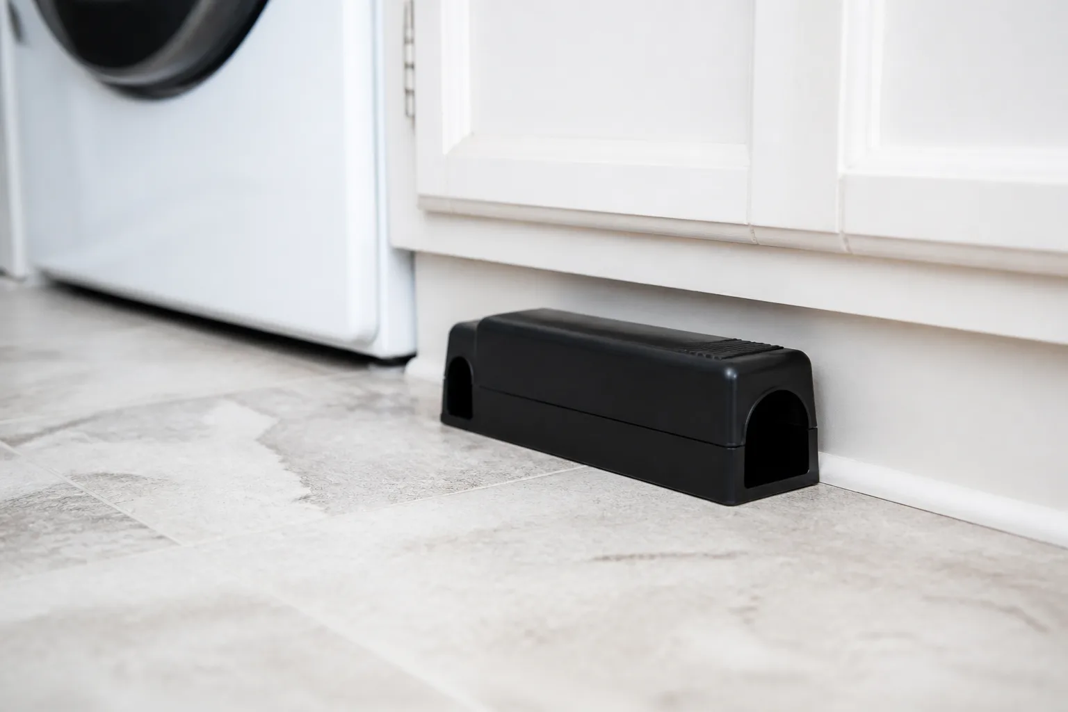 Black dual-entry covered snap trap on a laundry room floor against cabinets