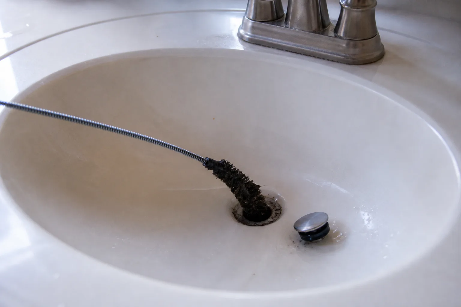 Stiff pipe brush scraping dark organic biofilm from a bathroom sink drain opening to remove drain fly larvae