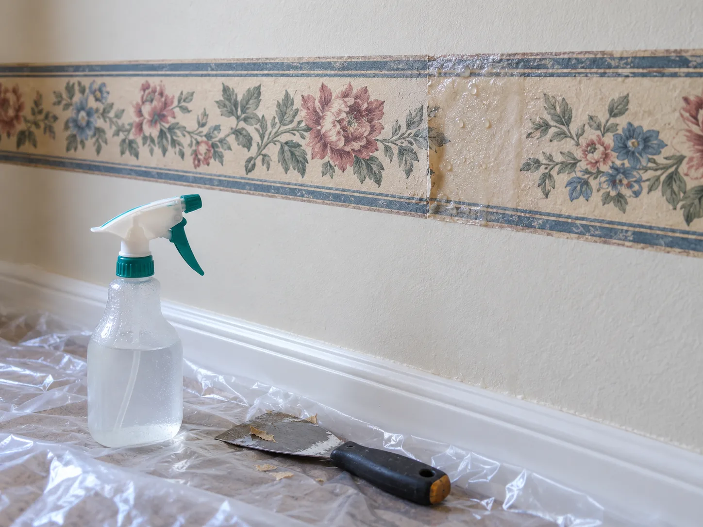 A small section of wallpaper border lightly dampened above a protected floor, with a scraper nearby