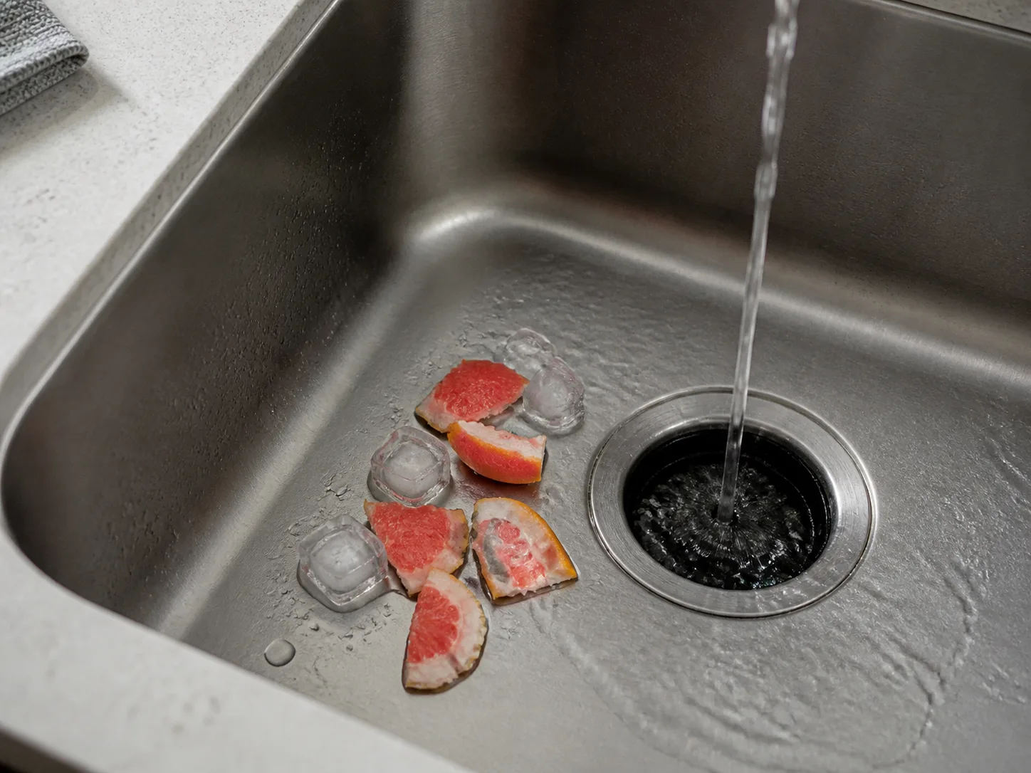 A few small grapefruit peel pieces and ice cubes beside a sink disposal under running cold water