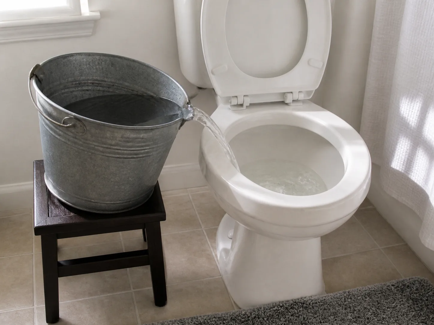 Bucket of reused dish-rinse water being poured into a toilet bowl for a manual flush