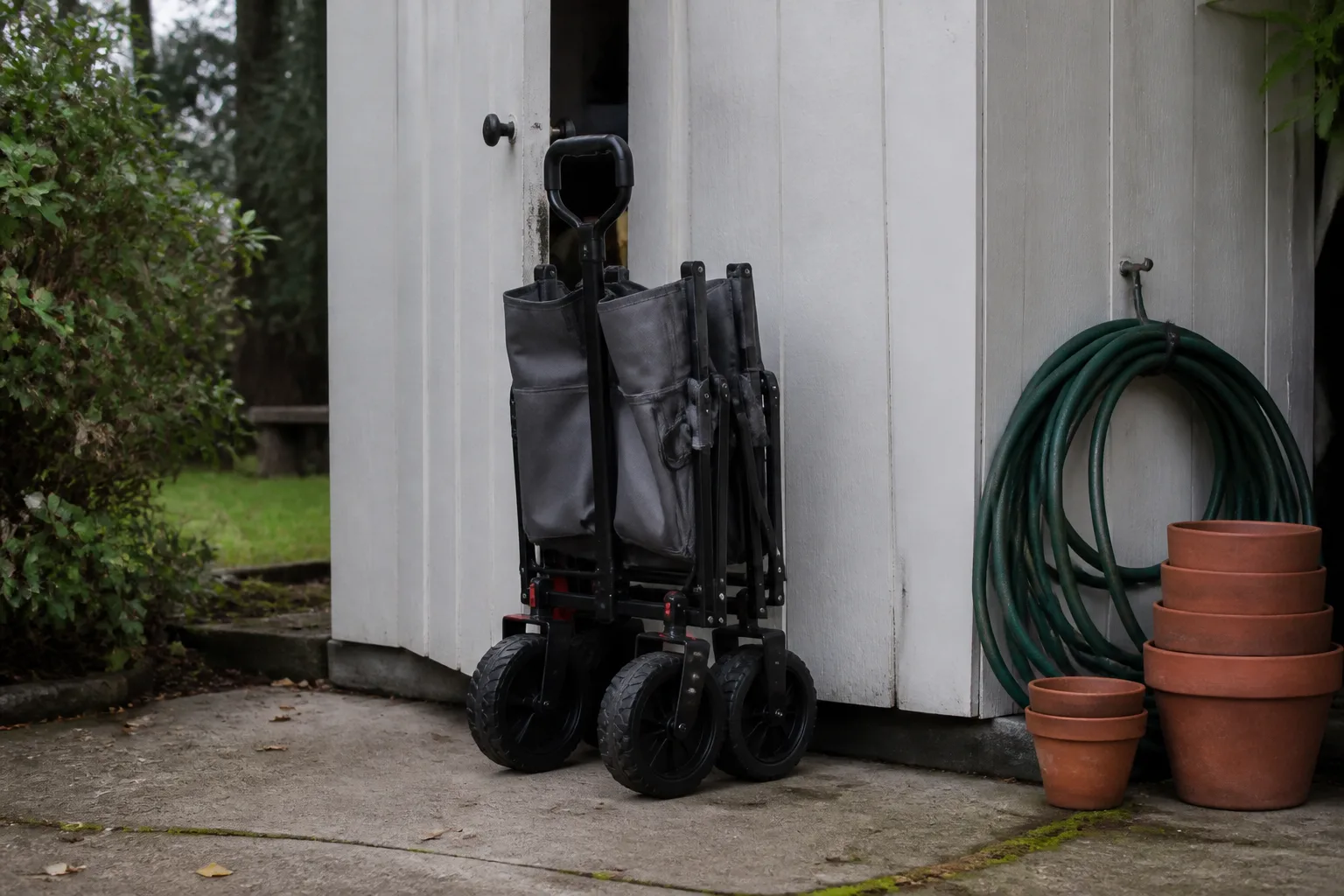 Folded fabric collapsible garden wagon with balloon tires beside a narrow shed on a patio